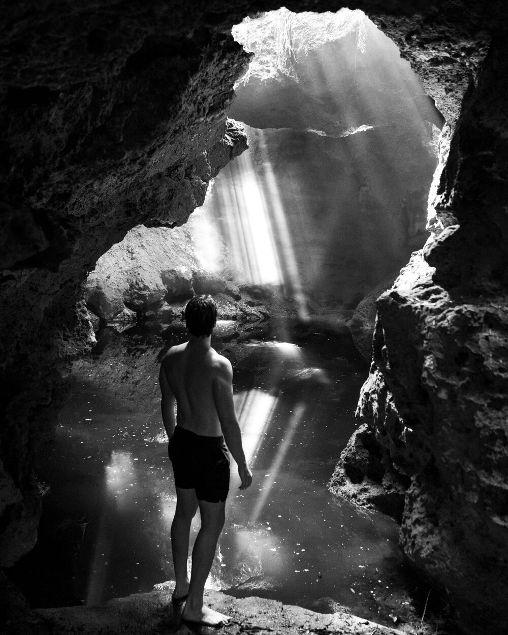 Brandon standing at the edge of a cave spring as light beams fall through, black and white